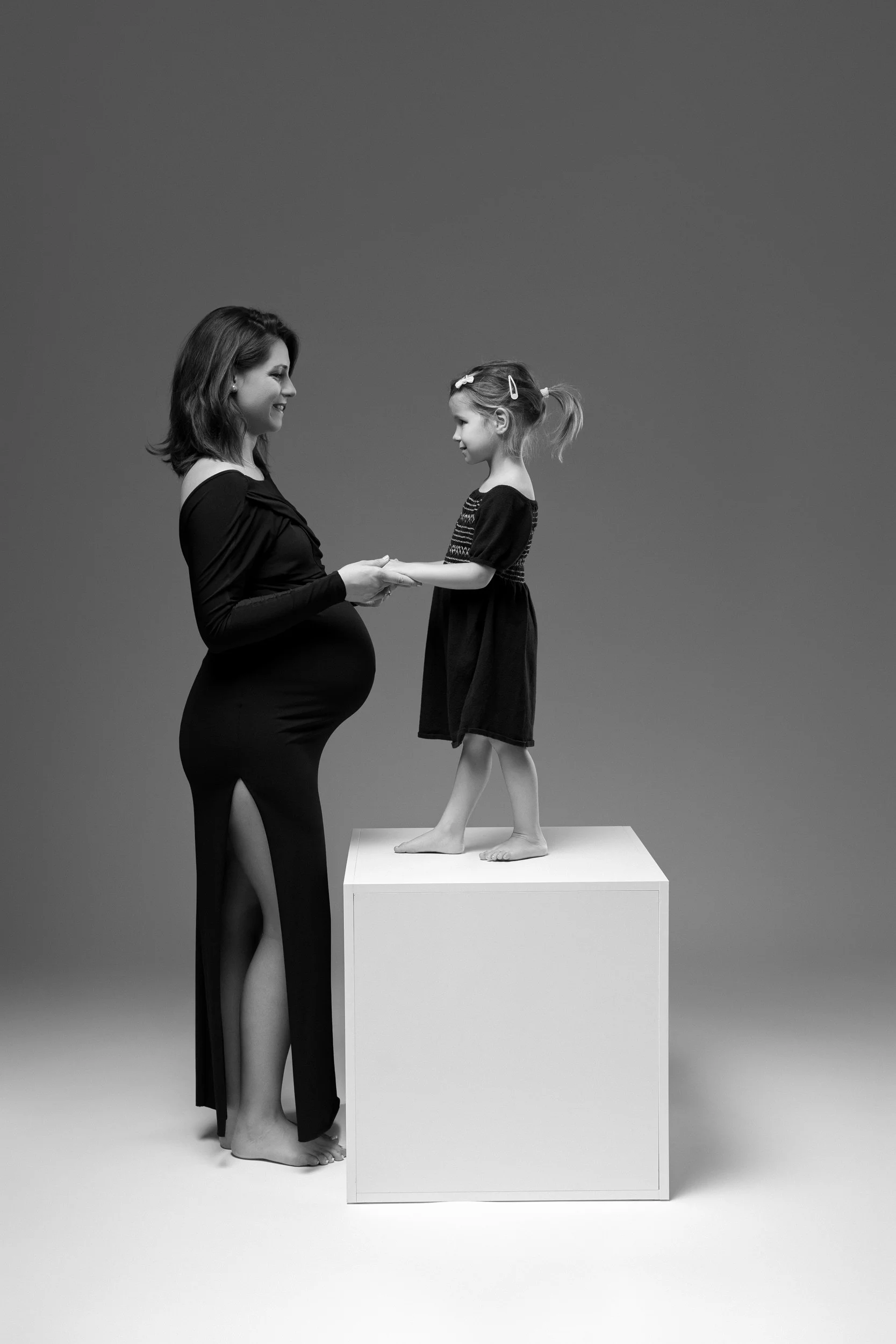 Noseča ženska in mlado dekle stojita na belem podstavku v črno-beli fotografiji. Ženska nosi črno obleko s stranskim razporkom, dekle pa črno obleko z mašno na hrbtu. Ozadje je enobarvno sivo.