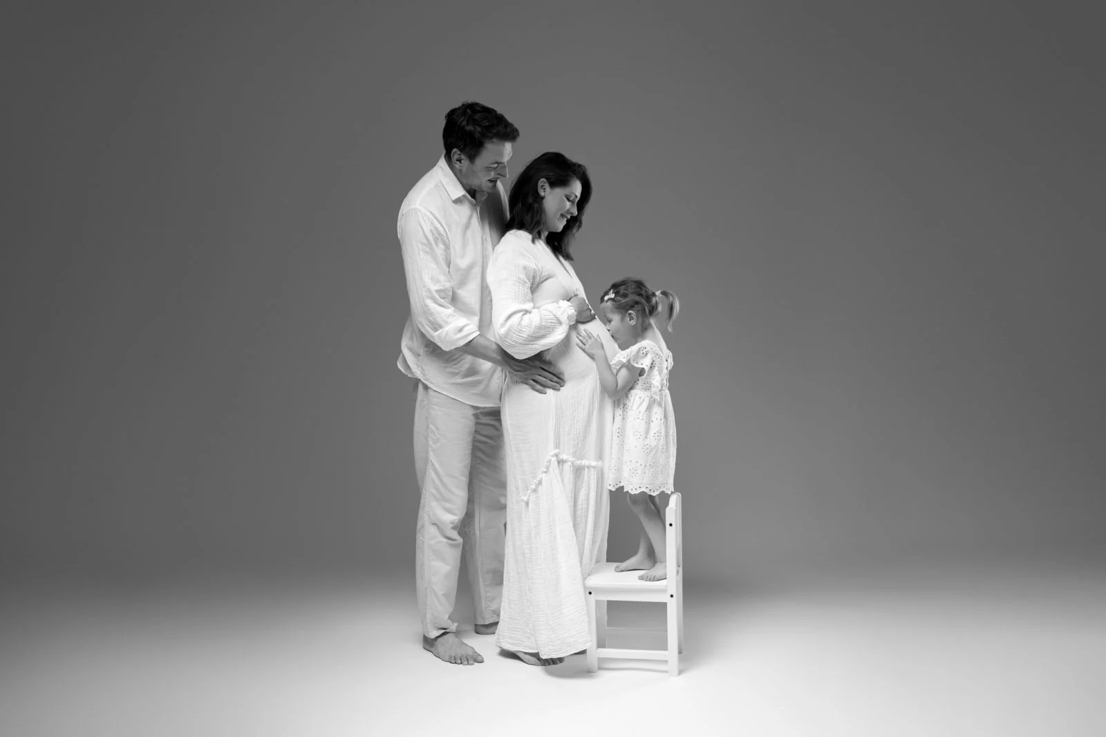 Družina treh v črno-beli fotografiji. Oblečeni so v bela oblačila. Oče drži mamo v naročju, hčerka pa stoji na belem stolčku.
