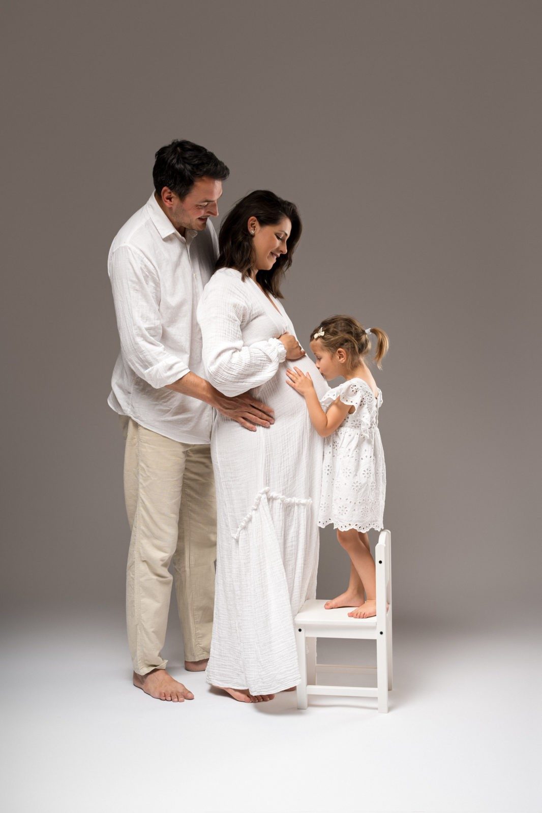 Družina treh stoji pred sivim ozadjem v studiu. Oče objema mamo in hčerko, mama drži belo tablo, hčerka stoji na belem stolčku.