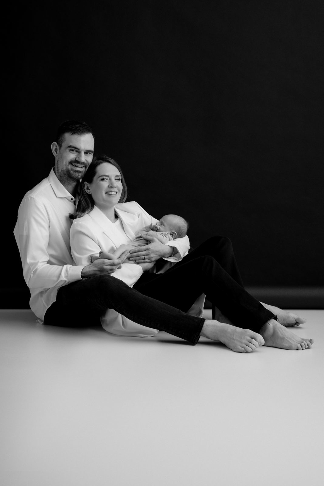 Črno-bela fotografija, ki prikazuje par, ki drži novorojenčka. Par sedi na belem tleh s črnim ozadjem. Moški nosi belo srajco in črne hlače, ženska pa belo bluzo in črne hlače. Novorojenček nosi belo pajacko in ga drži ženska.