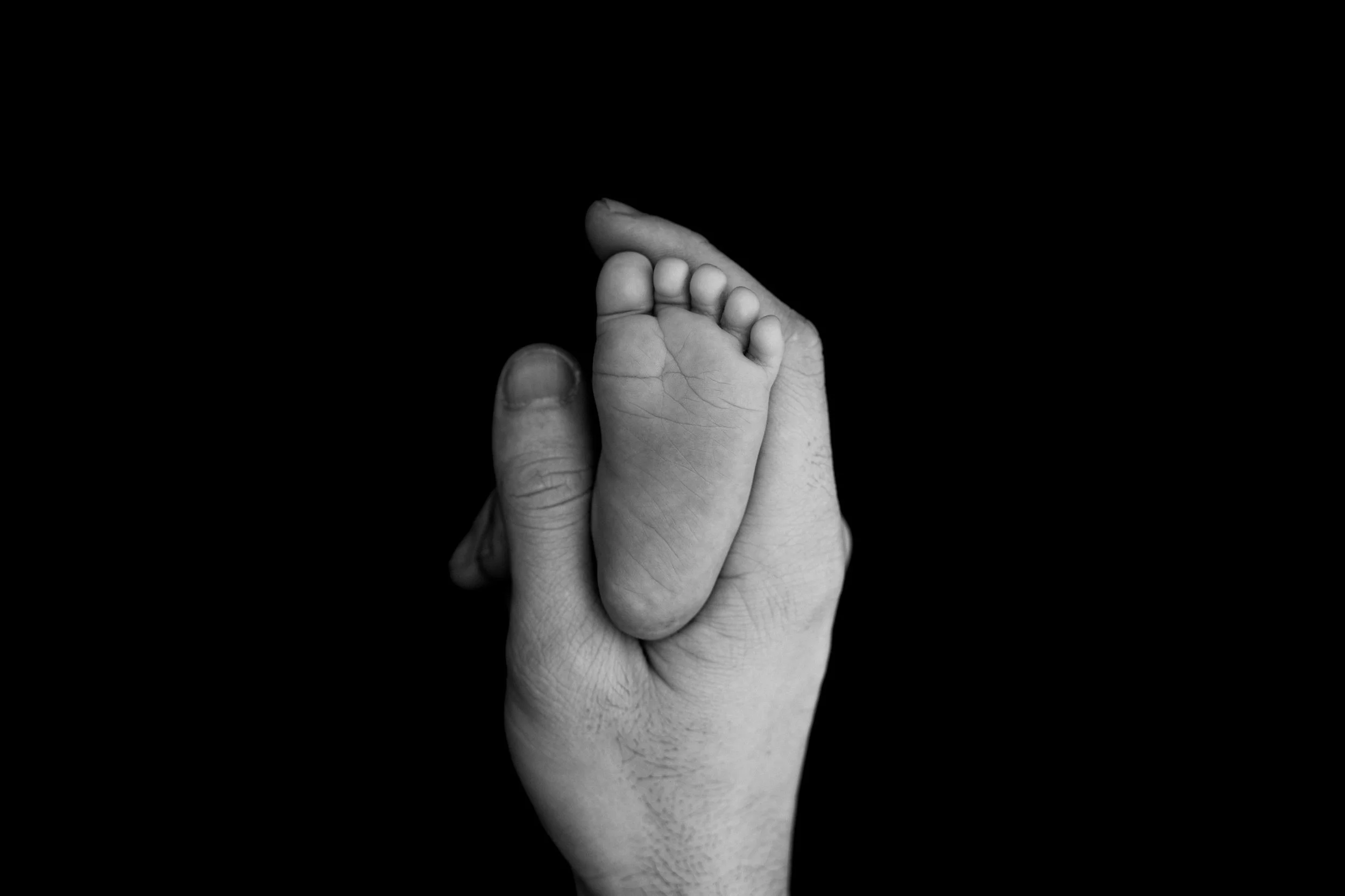 Nežen trenutek, ujet v črno-beli, ko otroška noga počiva na odrasli roki, ki poudarja razliko v merilu in teksturi.